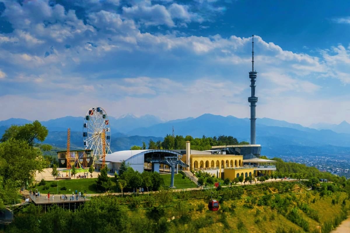 Almaty amusement park with Ferris wheel, yellow building, TV tower, and snow-capped mountains in background
