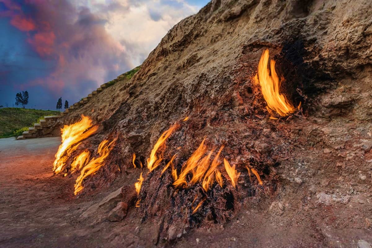 Natural gas flames burning on rocky hillside in Yanar Dag, Azerbaijan, with green vegetation and cloudy sky above