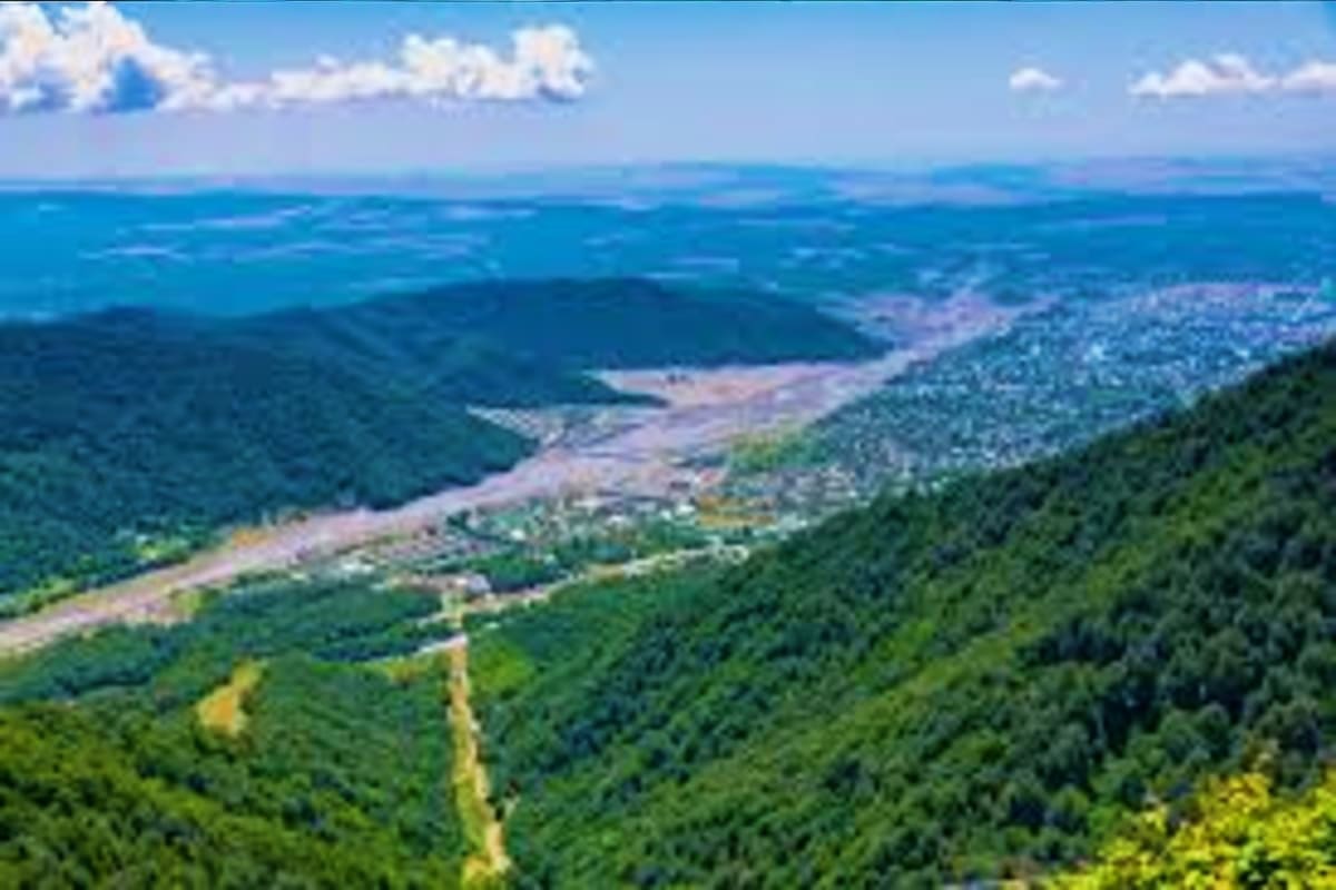 Aerial view of a winding river through dense green forest with mountains and blue sky with white clouds