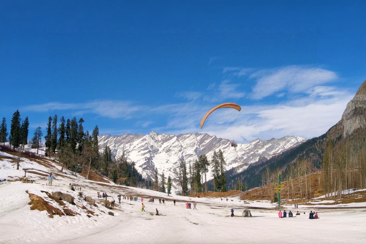 Paraglider soaring over snow-covered Solang Valley with snow-capped Himalayan peaks, pine forests, and tourists enjoying wint
