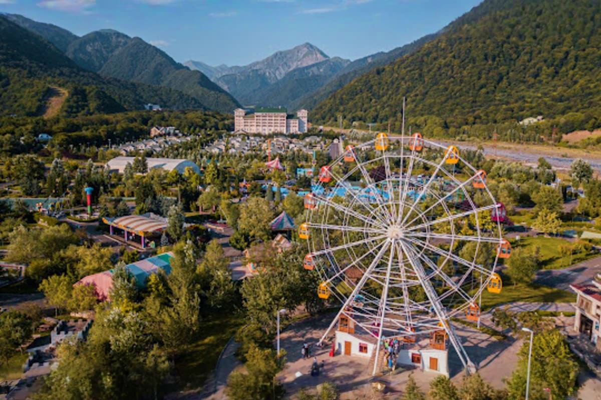 Aerial view of amusement park with colorful Ferris wheel surrounded by forested mountains and hotel building in valley landsc