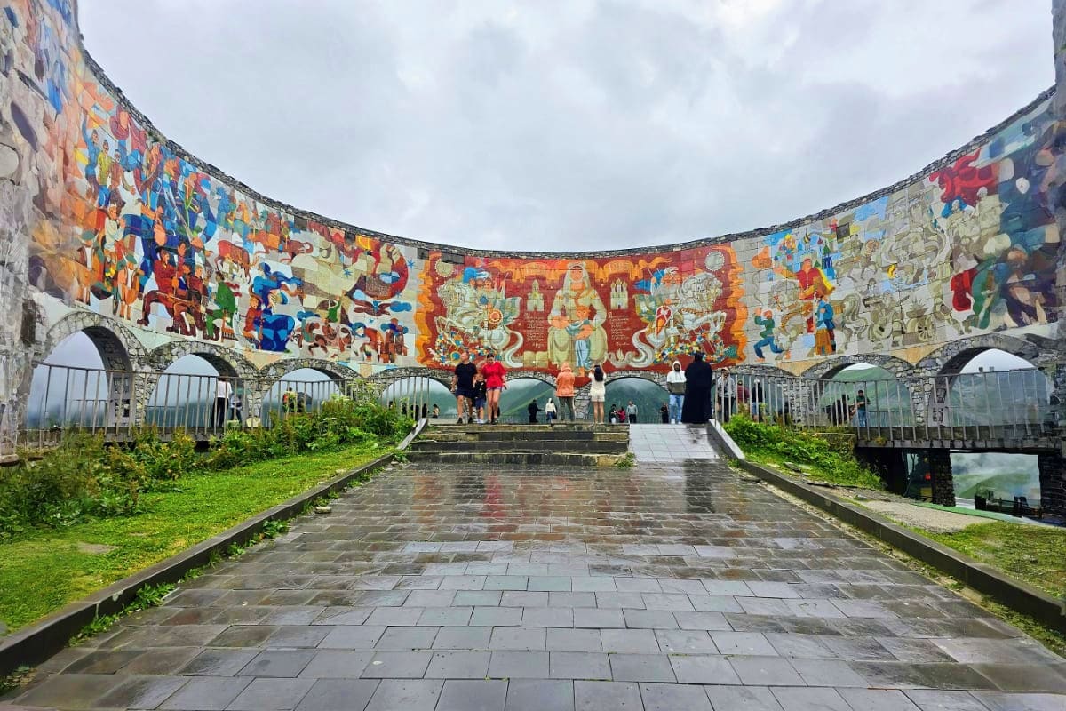 Circular courtyard with colorful murals depicting historical scenes, stone arches, and visitors walking on wet paved ground u