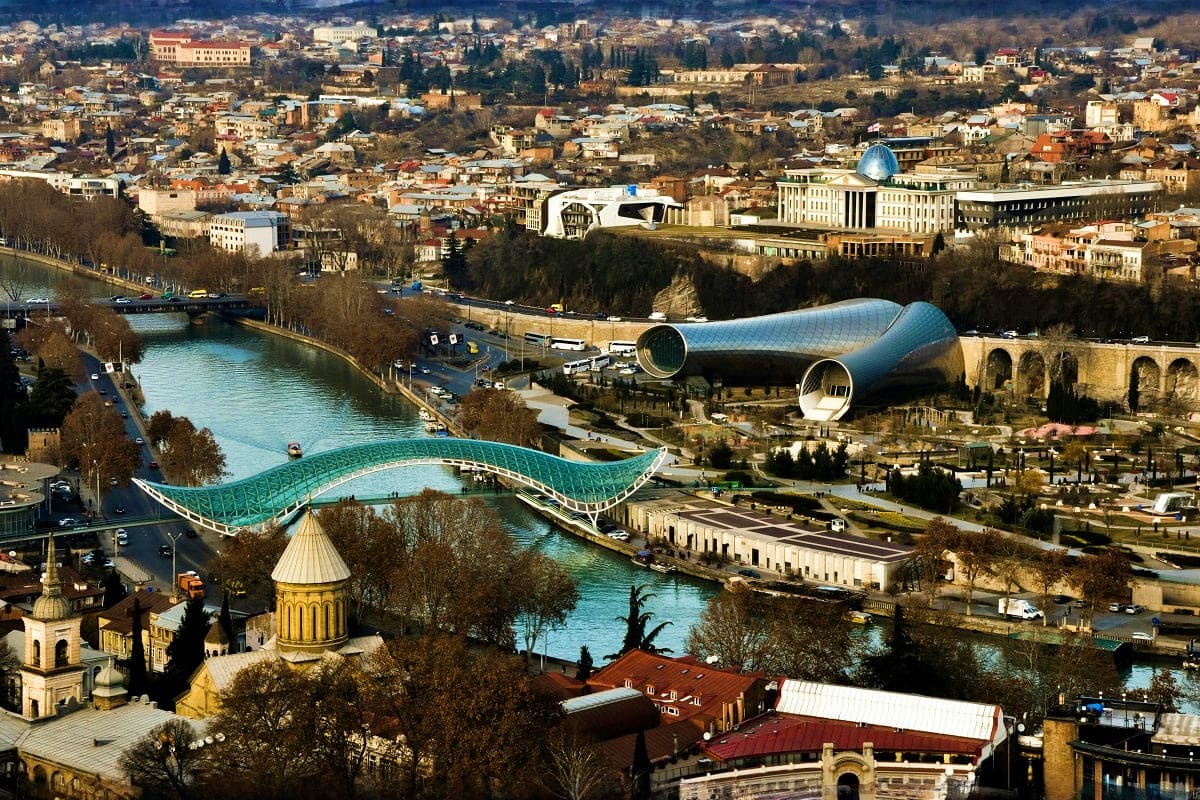 Aerial view of Tbilisi with turquoise Mtkvari River, modern wavy bridge, historic golden dome church, and contemporary archit