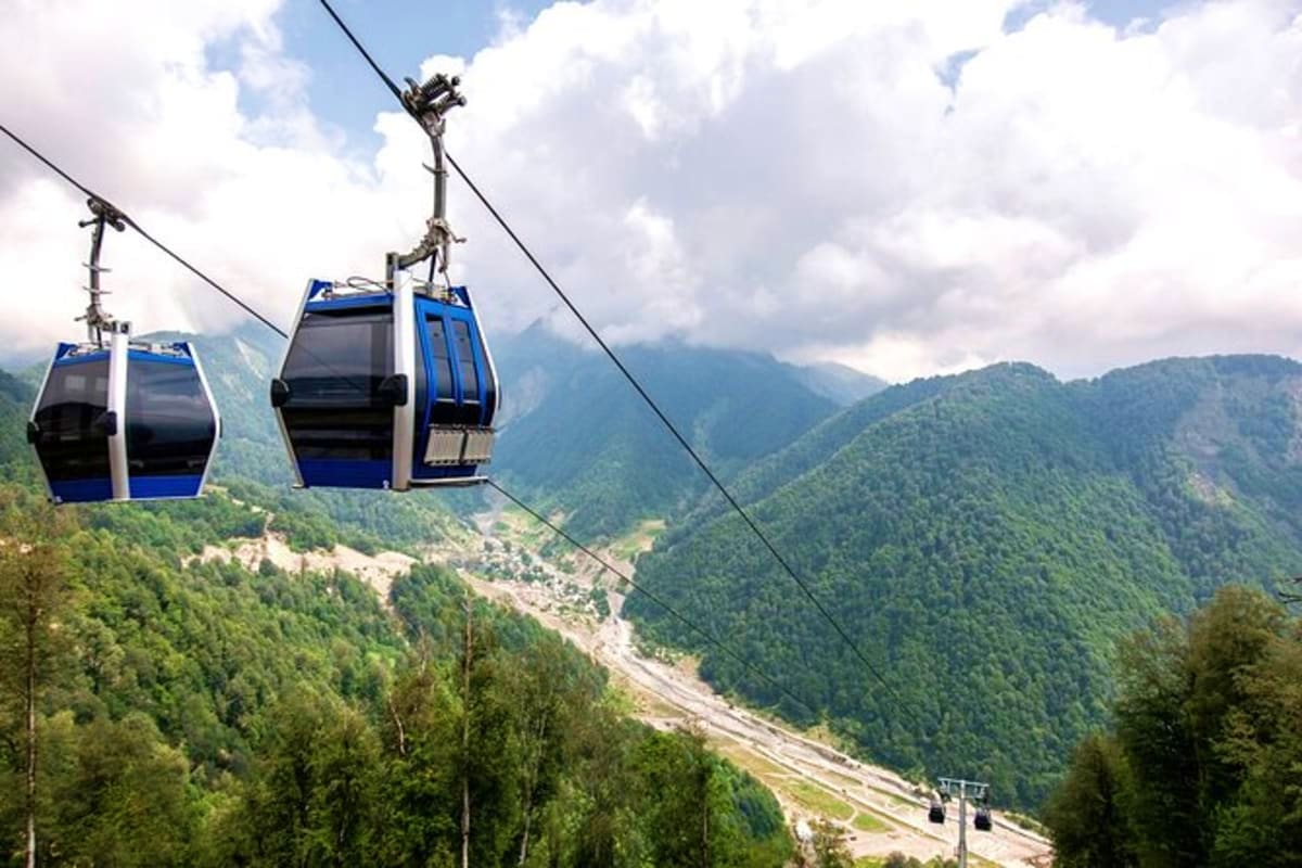 Cable car with blue and black cabins suspended over mountainous valley with forested slopes and river below