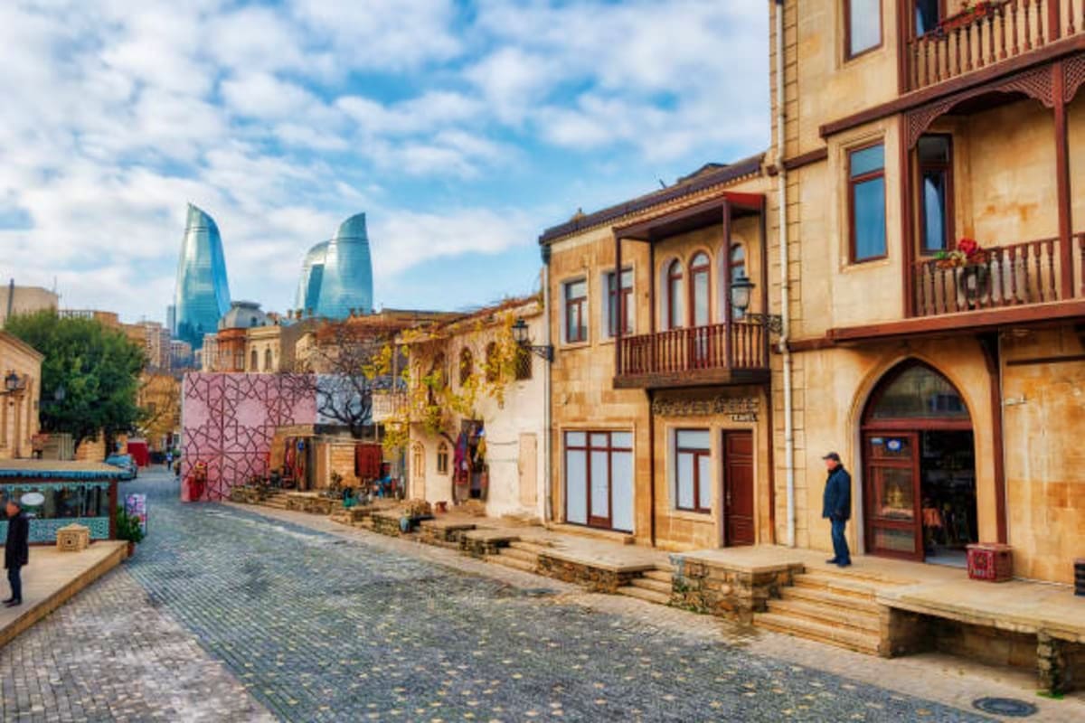 Historic stone buildings with wooden balconies on cobblestone street in Baku's Old City with modern glass towers visible in b