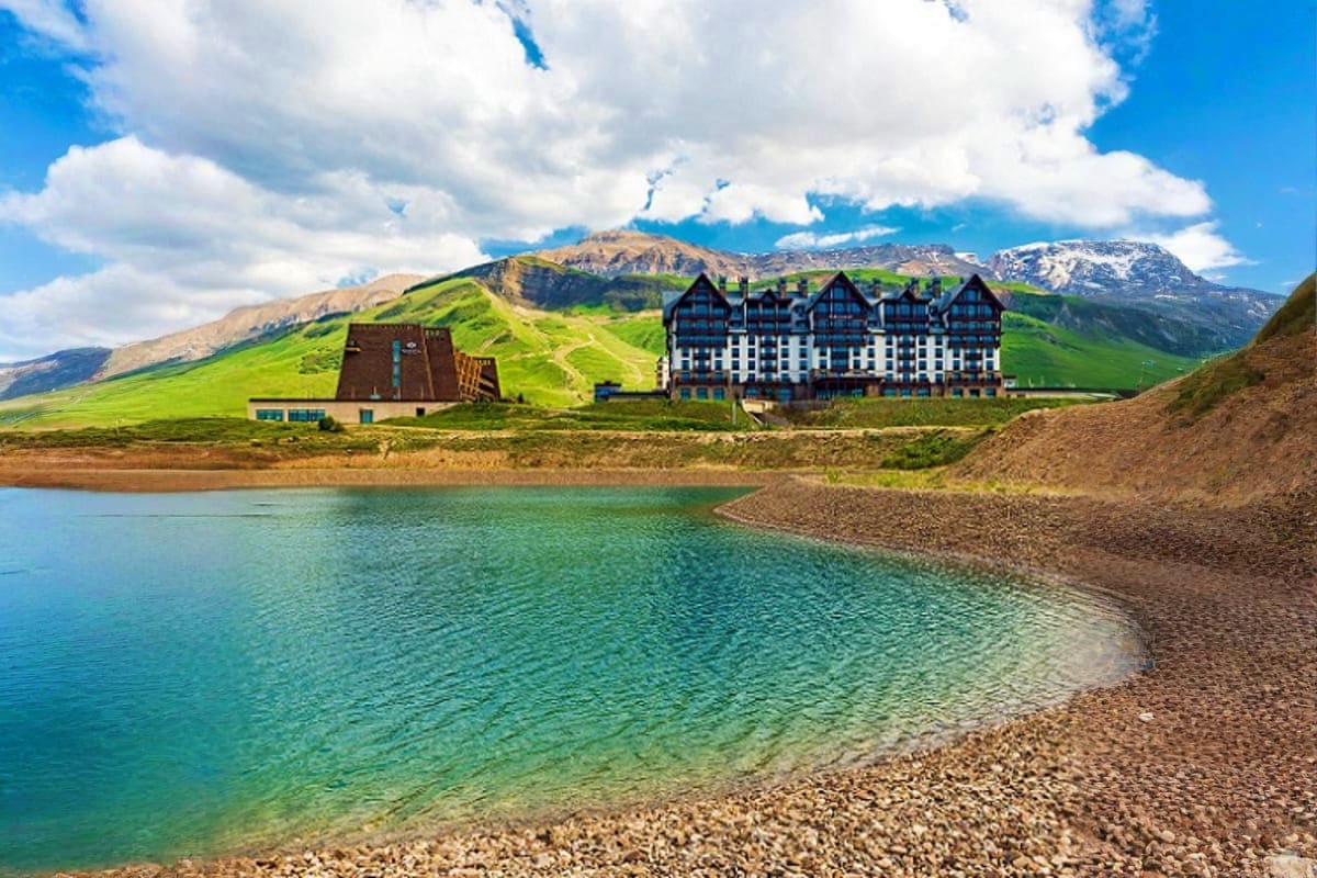 Mountain lodge and traditional building on emerald lake shore with snow-capped peaks and green slopes in Azerbaijan