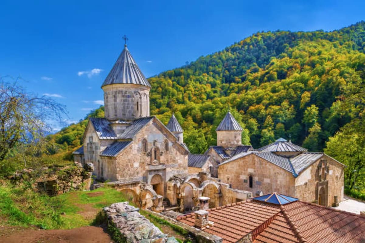 Historic Armenian monastery complex with stone buildings and conical domes nestled in forested mountains under blue sky