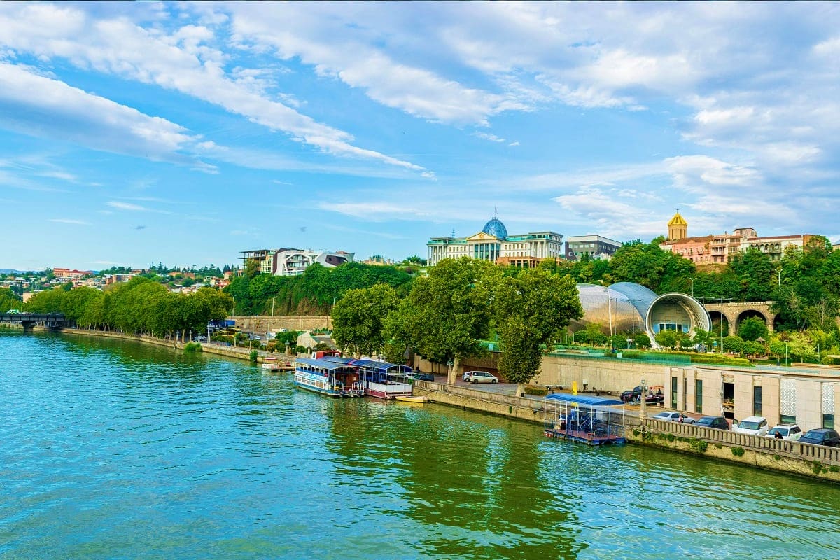 Tbilisi riverfront with Mtkvari River, tour boats, and historic cityscape with government buildings and golden dome church un