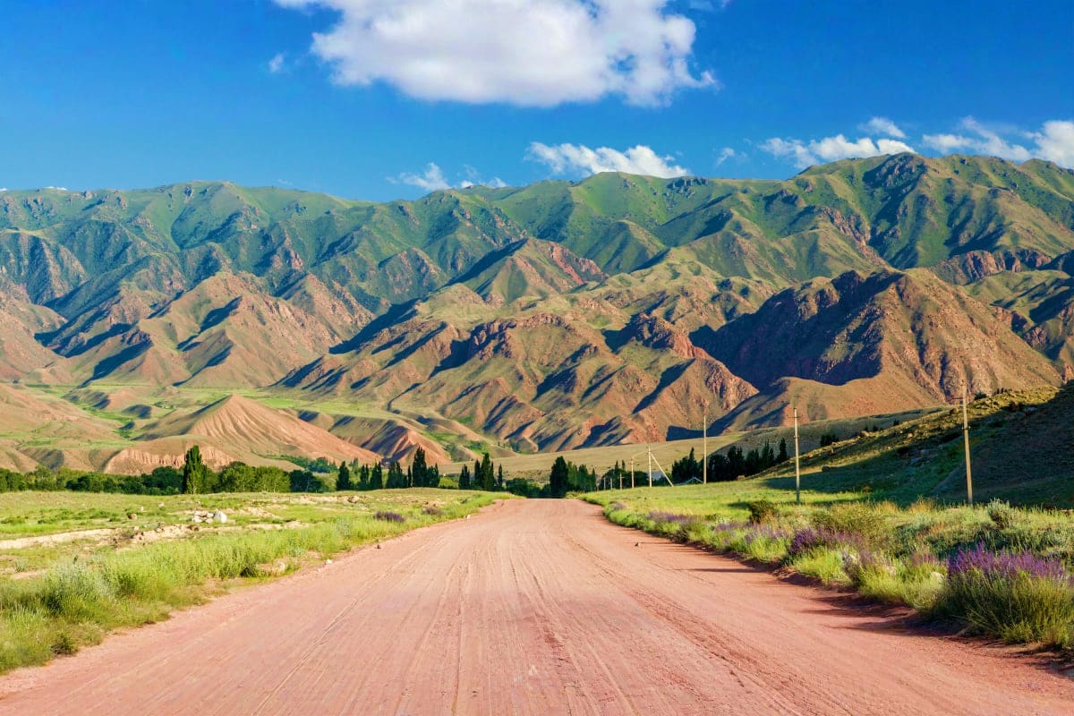 Classic Kyrgyzstan Unpaved red dirt road stretching through a valley toward dramatic, colorful mountains in Kyrgyzstan under blue sky