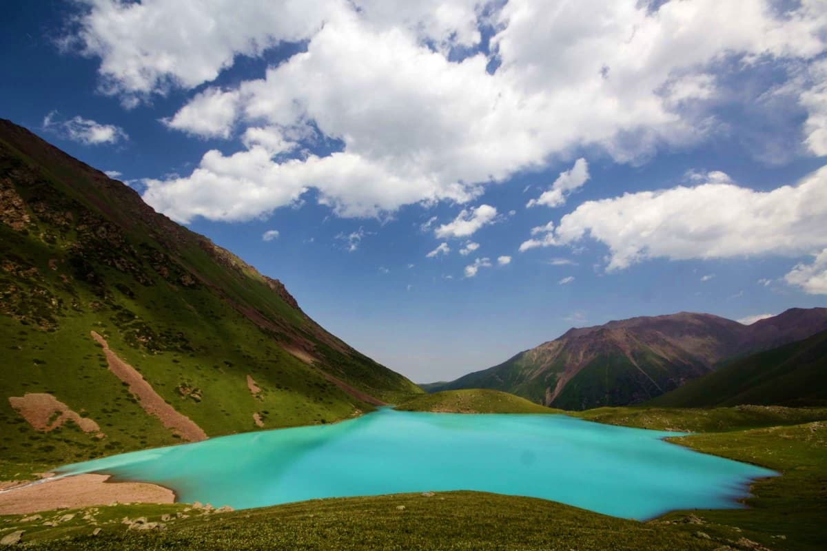 Classic Kyrgyzstan Turquoise alpine lake surrounded by green mountain slopes and rocky peaks under blue sky with white clouds in Kyrgyzstan
