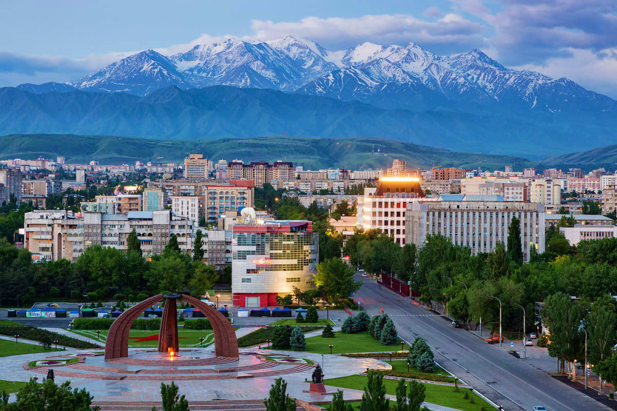 Classic Kyrgyzstan Almaty cityscape with snow-capped mountains in background, featuring Ala-Too Square monument and modern buildings at sunset