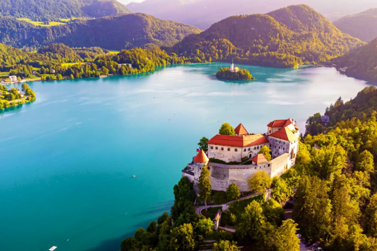 Bled Castle perched on a cliff overlooking turquoise Lake Bled surrounded by forested mountains and small island church in Sl