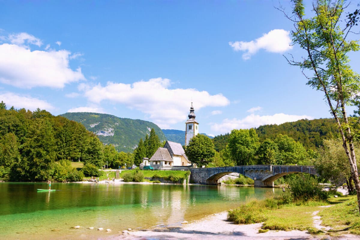 Church with bell tower reflected in emerald lake surrounded by forested mountains and stone bridge in Slovenia