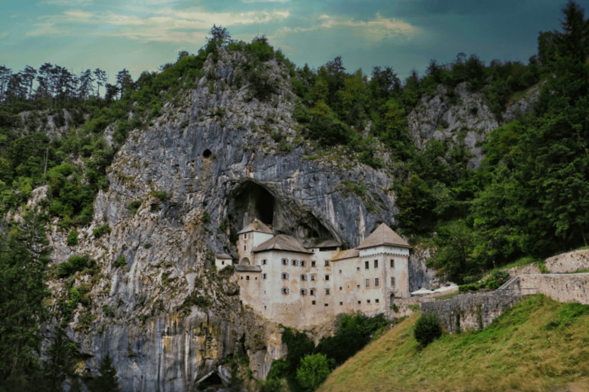 Predjama Castle nestled beneath a large cave opening in a limestone cliff, surrounded by green forests and stone fortificatio