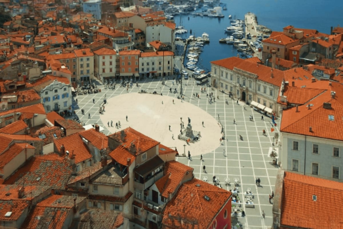 Aerial view of a coastal Mediterranean town square surrounded by terracotta roofs, with boats moored in the harbor beyond