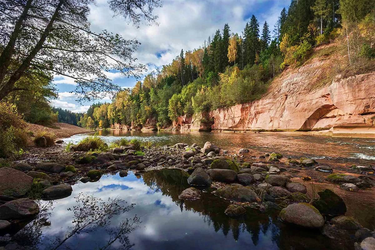 Scenic river valley with red sandstone cliffs, forested hillsides, and rocky shoreline reflecting blue sky and clouds in Latv