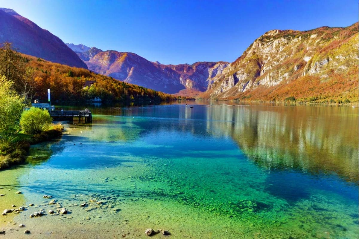 Crystal-clear alpine lake surrounded by autumn forest and rocky mountains with a small wooden boat dock on the shore