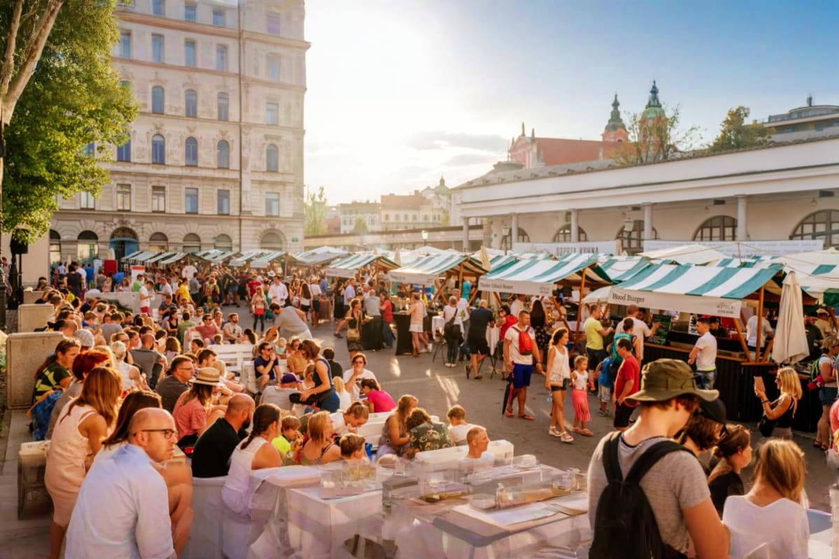 Busy outdoor market in Ljubljana's city square with white striped vendor tents, historic architecture, and church spires in b
