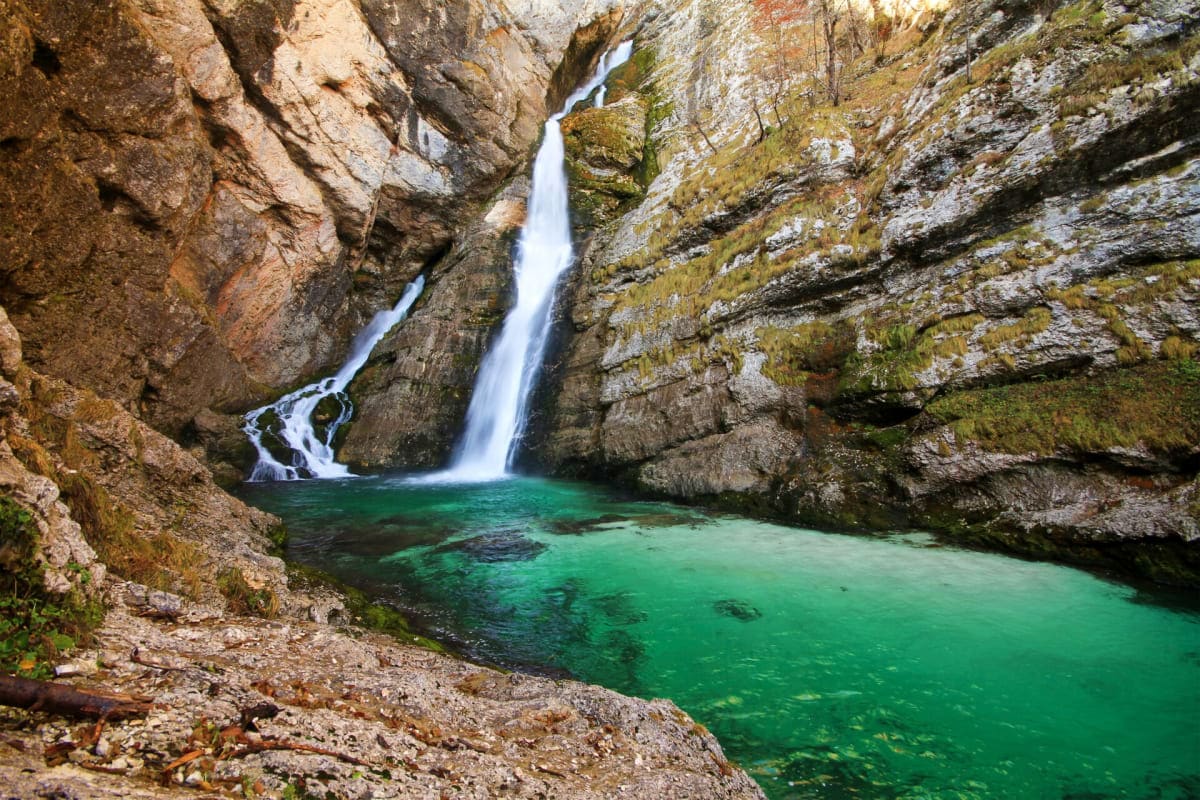 Waterfall cascading into emerald green pool surrounded by rocky canyon walls covered in moss in Alpine gorge