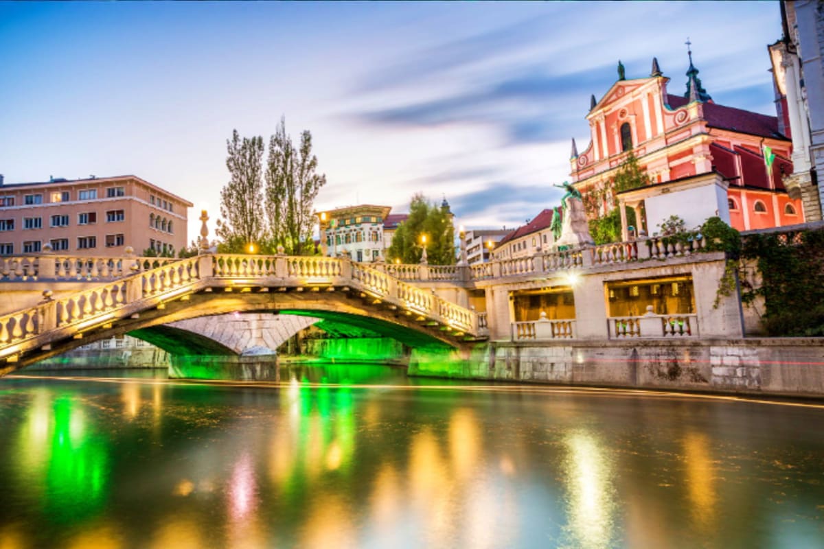 Illuminated stone bridge crossing green-lit river at dusk with red baroque church and historic buildings in Ljubljana, Sloven