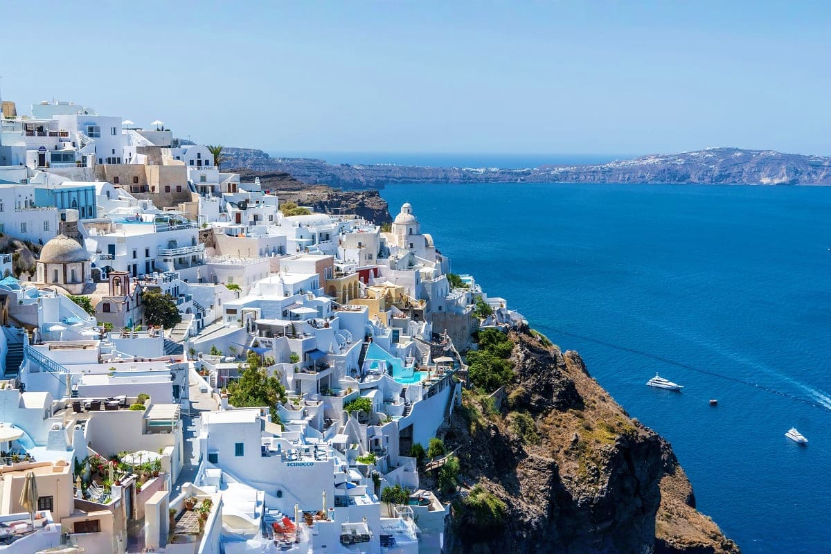White-washed buildings cascading down cliffs in Oia, Santorini, with blue Aegean Sea and sailboats below