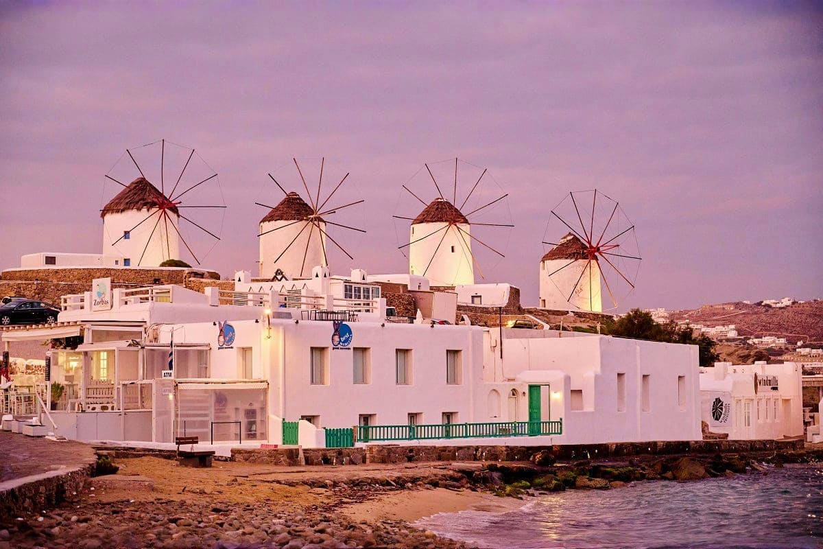 Traditional whitewashed buildings with green doors and iconic windmills at sunset in Mykonos, Greece