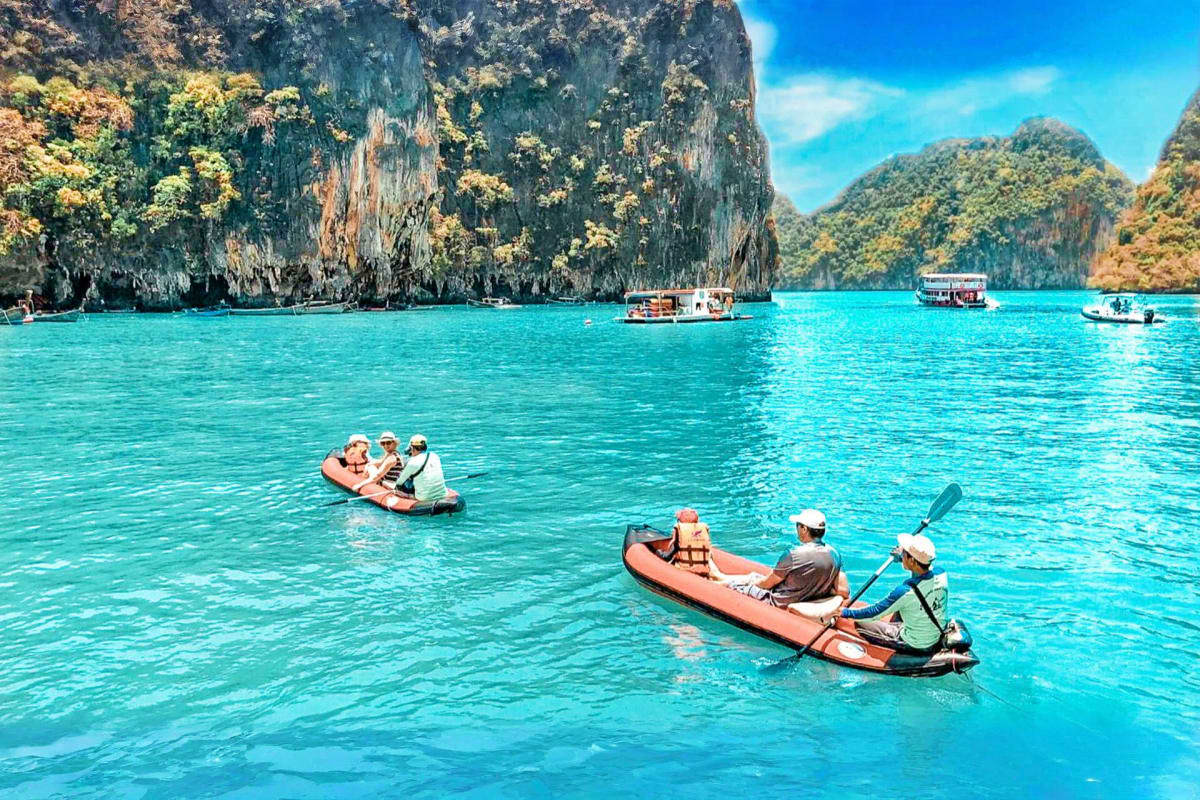 Tourists kayaking in turquoise water near limestone cliffs at Phang Nga Bay with tour boats in background