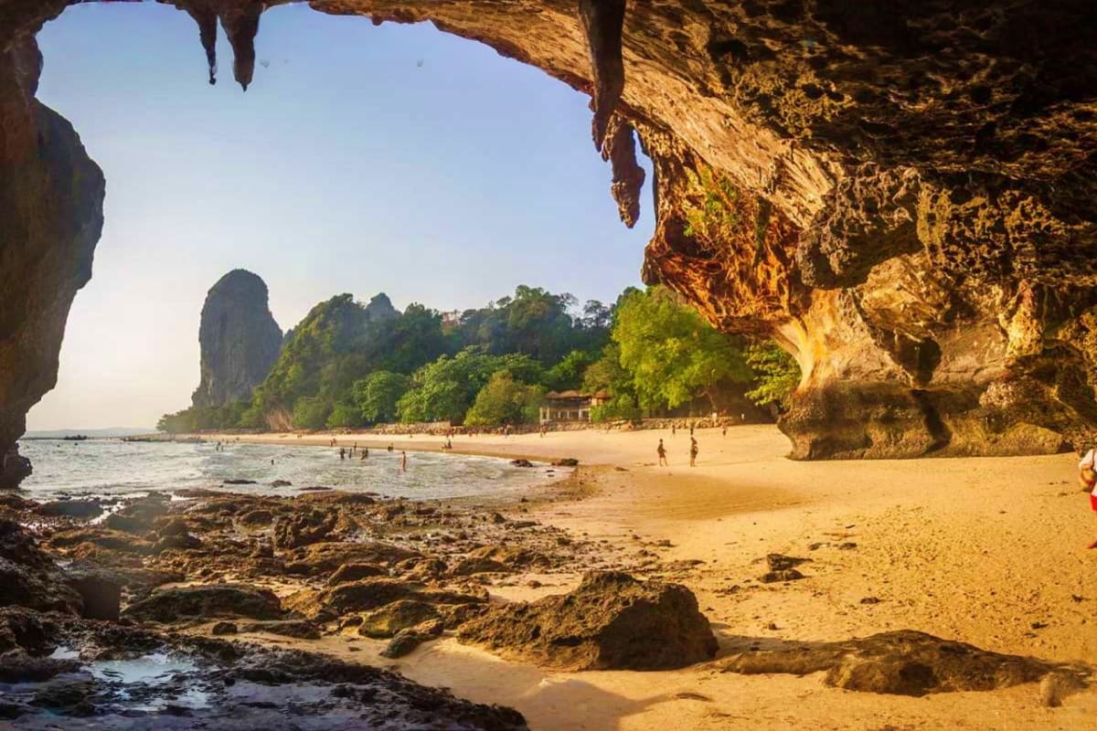 View from inside a cave of Railay Beach in Krabi with limestone cliffs, golden sand, turquoise water, and tourists swimming