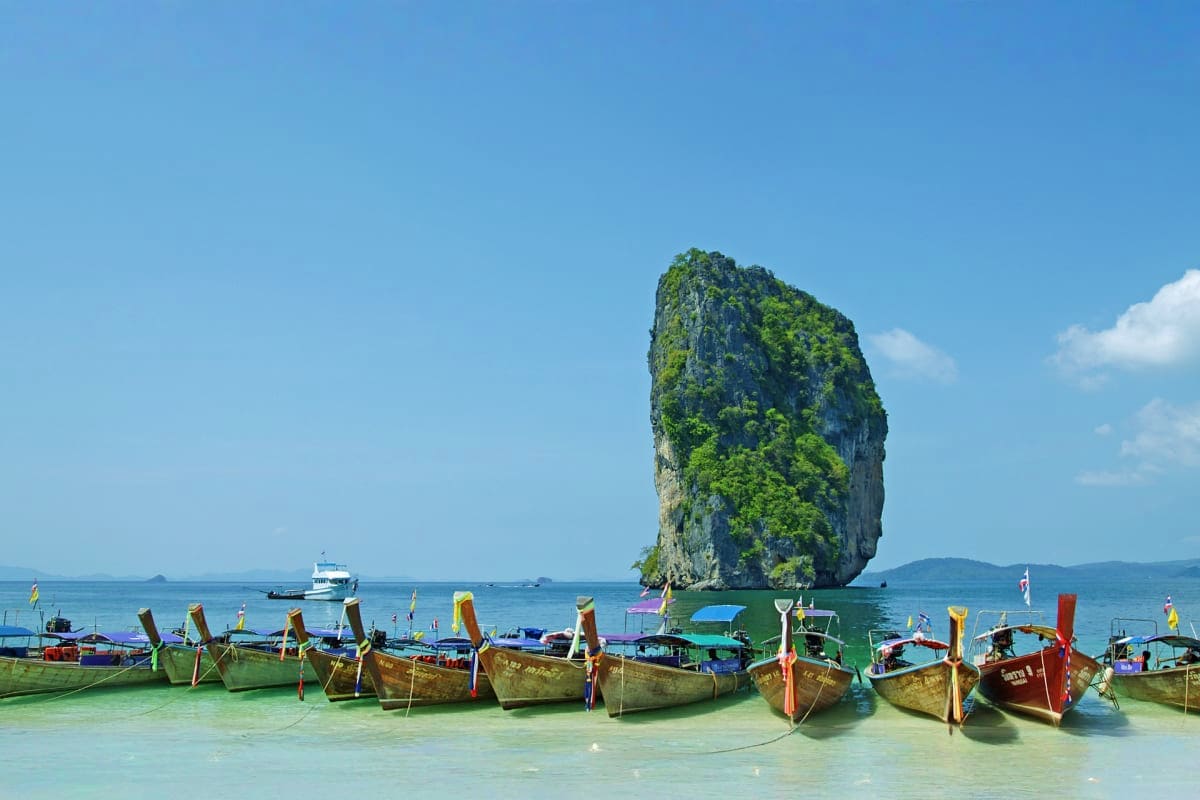 Traditional longtail boats in turquoise shallow water with dramatic limestone rock formation rising from the sea in Phuket, T