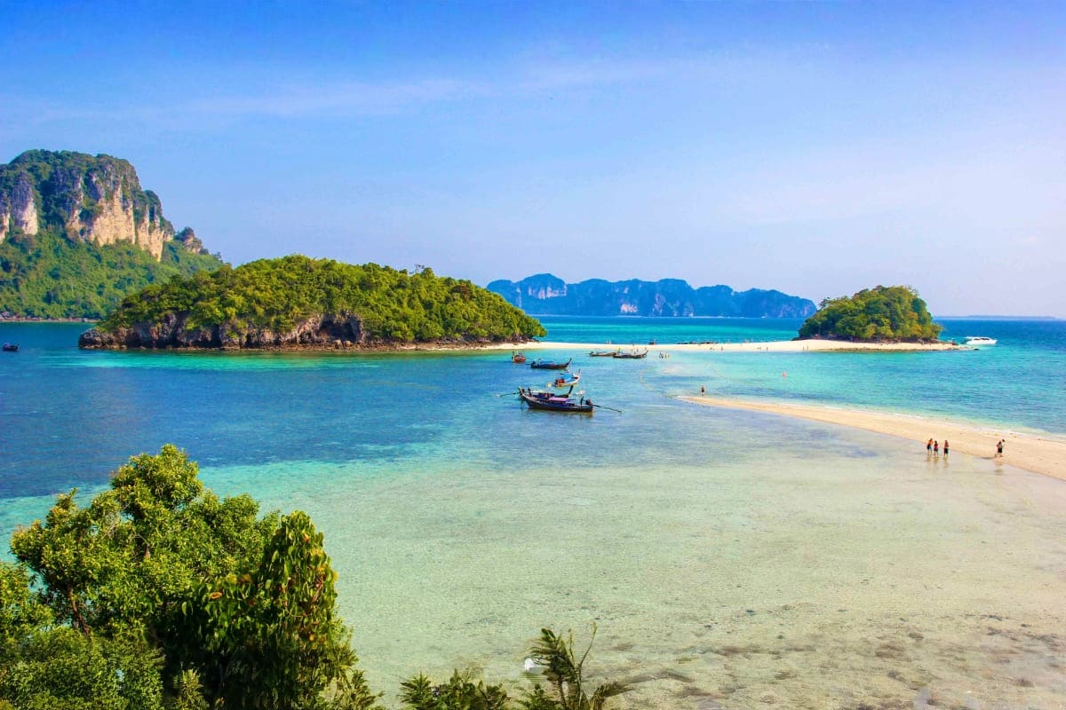 Tropical beach cove with turquoise water, limestone cliffs, traditional longtail boats, and white sand beach in Krabi, Thaila