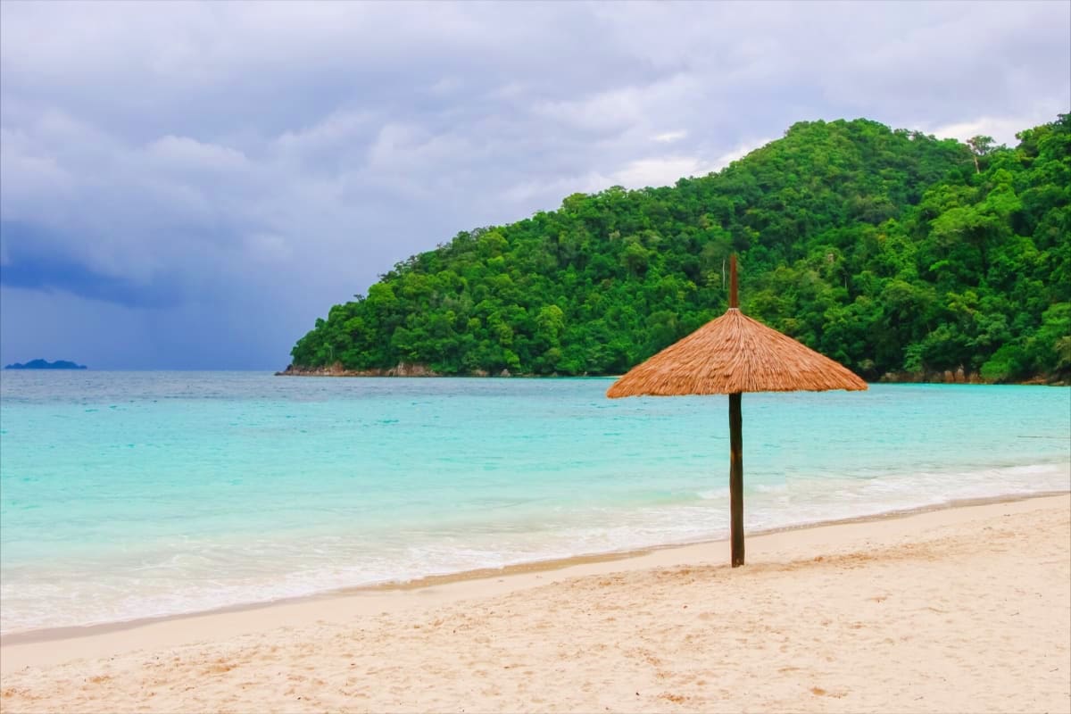 Tropical beach scene with thatched umbrella, turquoise water, white sand, and forested hillside in Andaman Islands