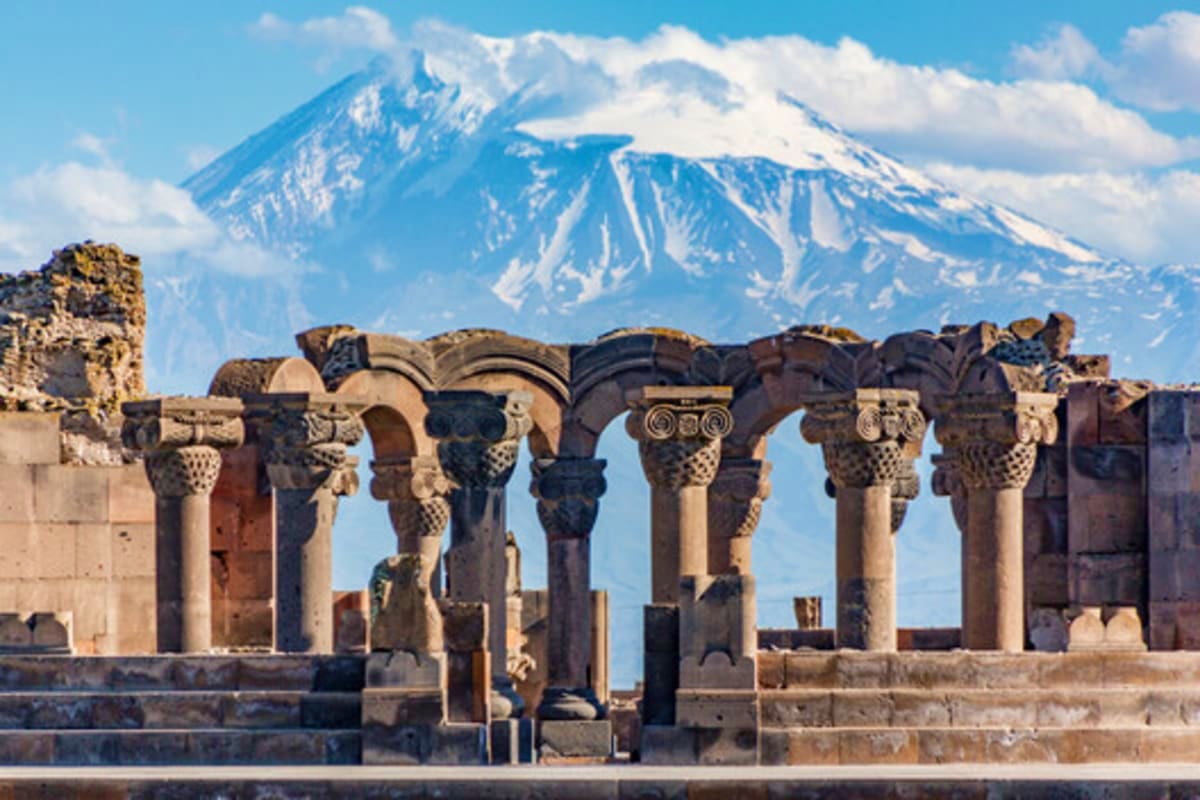 Ancient stone columns with ornate capitals at Persepolis ruins, snow-capped Mount Ararat in background under blue sky