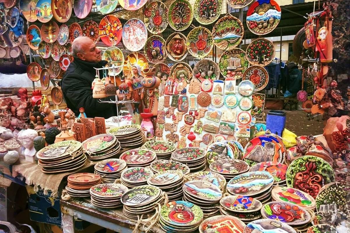 Vendor at Armenian souvenir market stall displaying colorful decorative plates, pottery, and traditional crafts