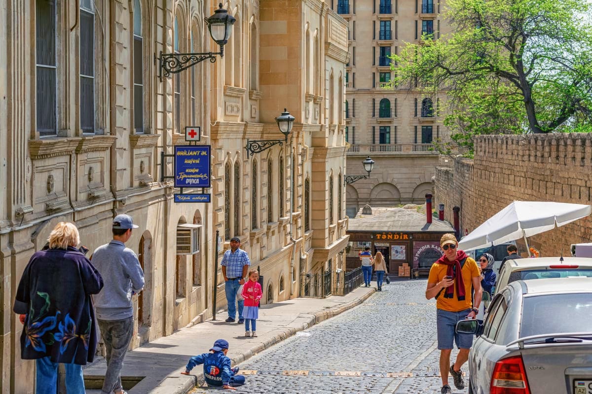 Historic cobblestone street in old Baku with traditional stone buildings, tourists, and ornate street lamps on a sunny day