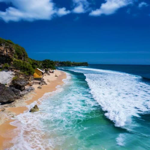 Scenic Bali beach with turquoise waves, white sand, rocky cliffs and tropical vegetation under clear blue sky