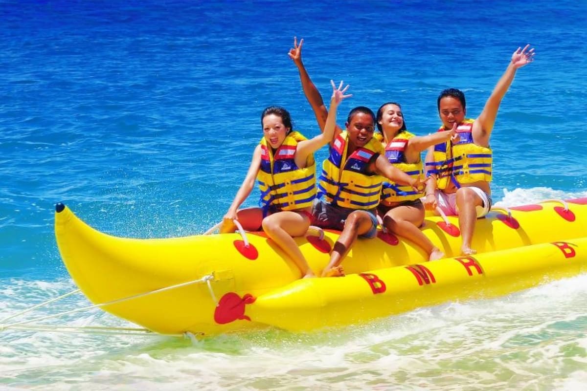 Bali Thrill & Serenity Group of five people riding a yellow banana boat in crystal blue ocean water with their arms raised in excitement