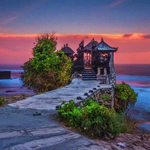 Ancient Hindu temple at sunset on rocky Bali coastline with dramatic purple and orange sky, surrounded by lush green vegetati