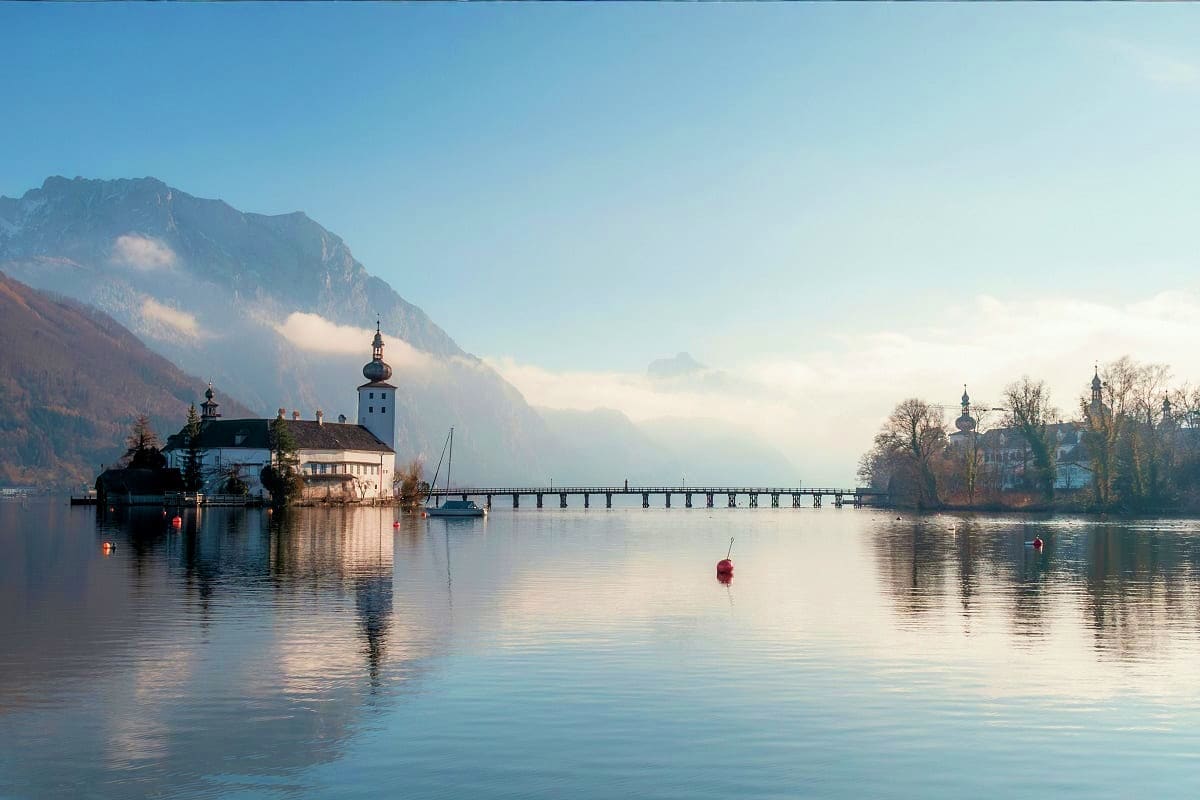 Scenic Alpine lake with white church and castle reflected in calm water, surrounded by mountains and wooden footbridge in mor