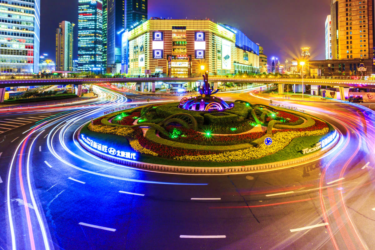 Night cityscape of Baku with illuminated roundabout featuring colorful flower gardens, light trails from traffic, and modern 