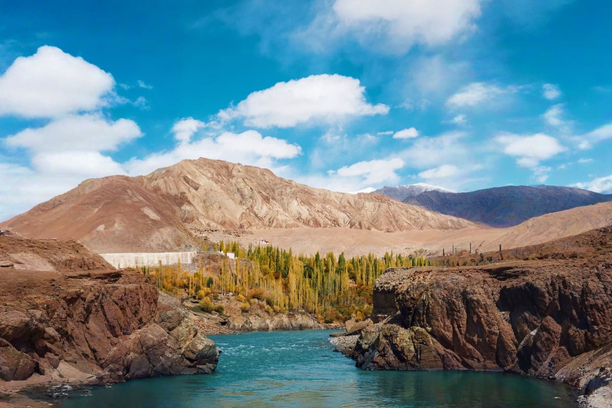 Turquoise river flowing through rocky canyon with poplar trees and snow-capped mountains under blue sky in Azerbaijan