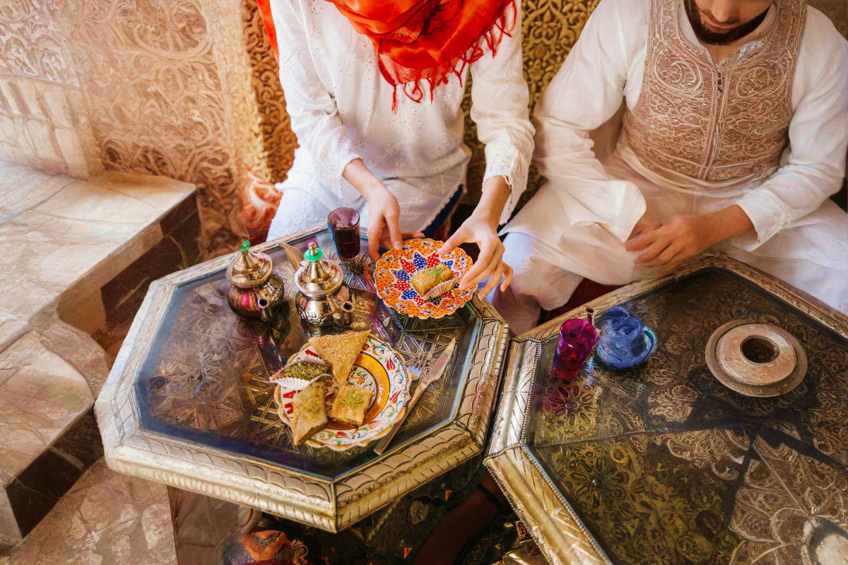 People in traditional white clothing sharing food and tea at an ornate table with Moroccan-style décor and pastries