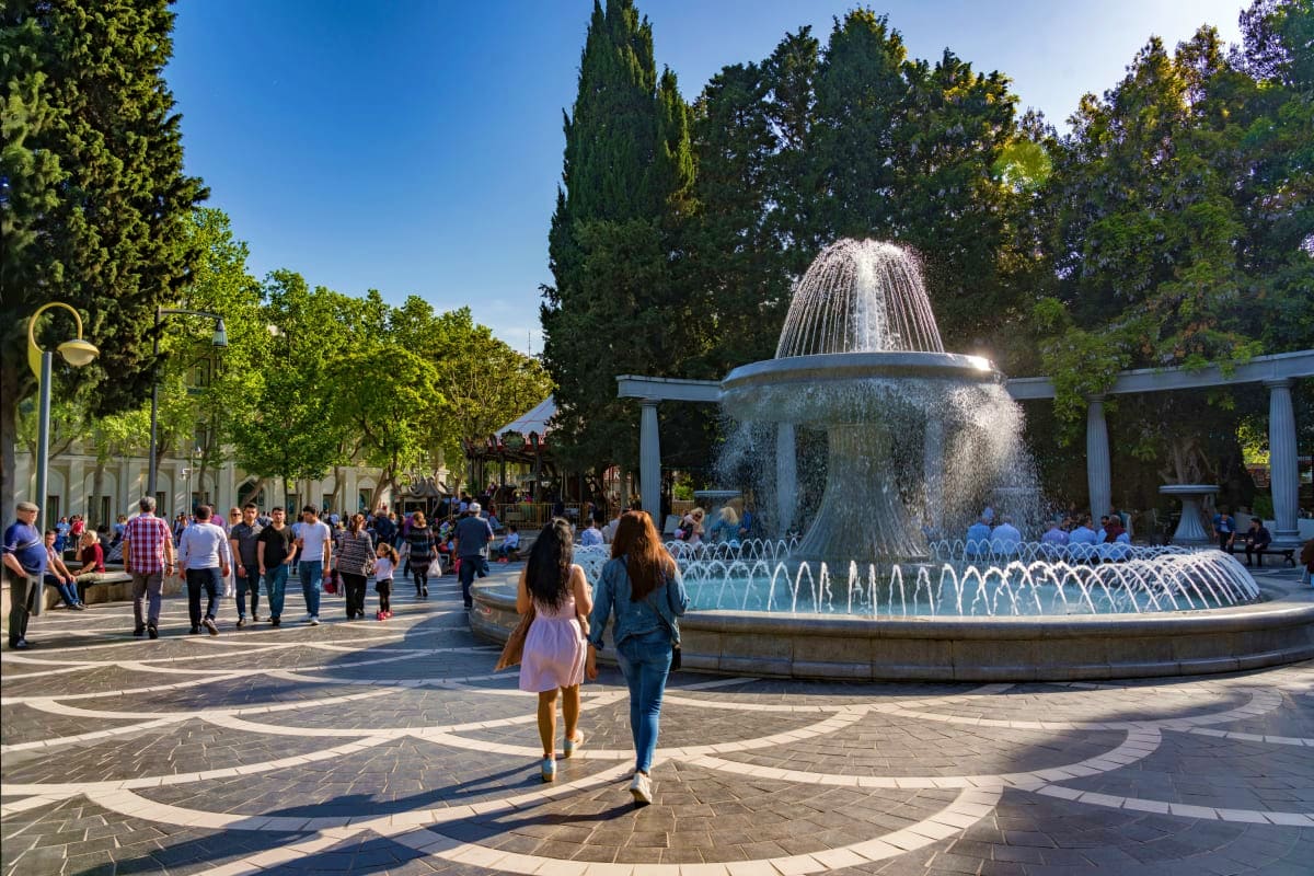 Visitors strolling around a grand tiered fountain in a public square surrounded by cypress trees and classical columns under 