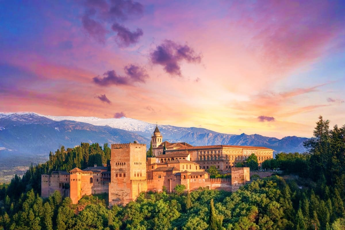 Alhambra palace in Granada at sunset with snow-capped Sierra Nevada mountains in background and green forests in foreground