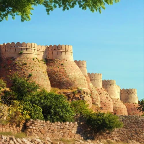 Ancient sandstone fort with crenellated walls perched on rocky hillside, surrounded by green vegetation under clear blue sky
