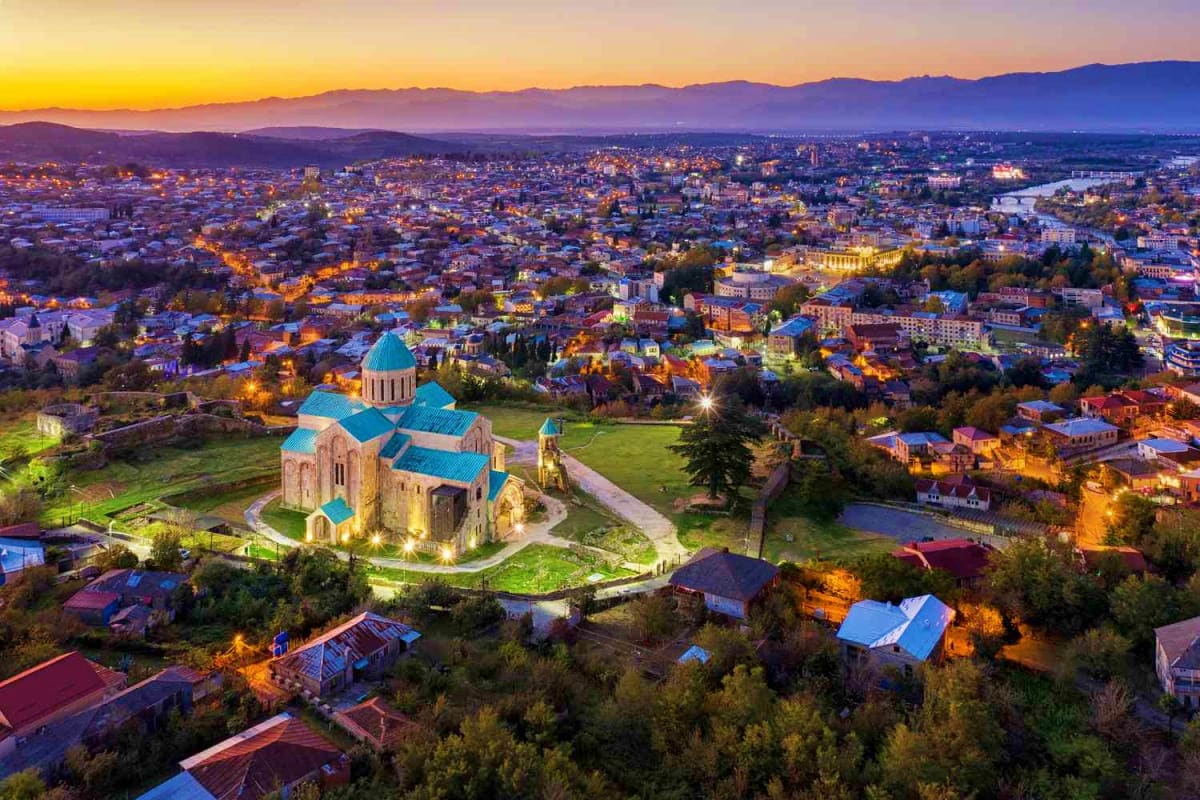 Aerial view of Tbilisi at twilight with illuminated Armenian church featuring blue dome in foreground and city lights spreadi