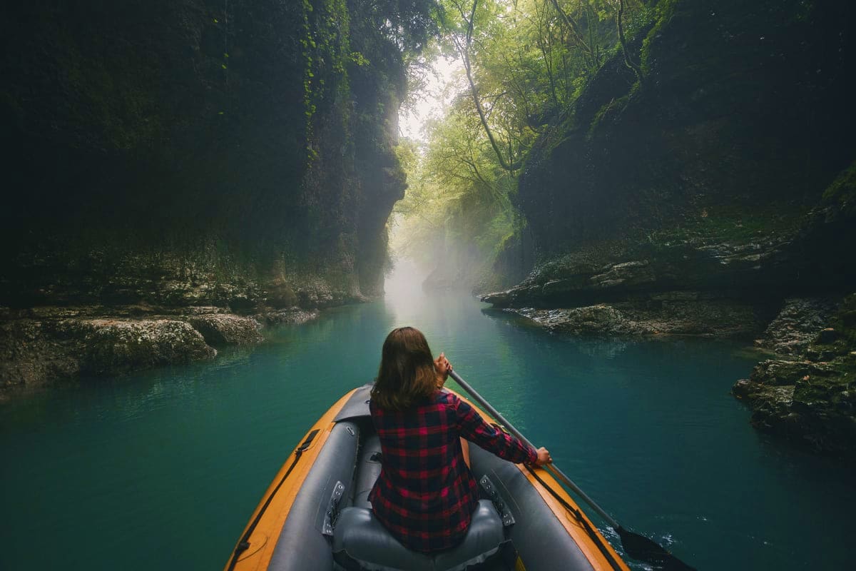 Woman paddling inflatable boat through misty turquoise river canyon surrounded by towering moss-covered limestone cliffs