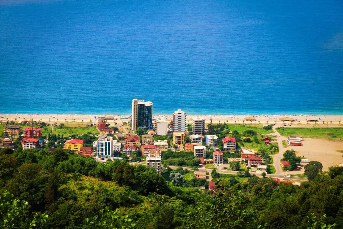 Aerial view of a Georgian Black Sea coastal town with hotels, sandy beach, turquoise water, and lush green forest surrounding