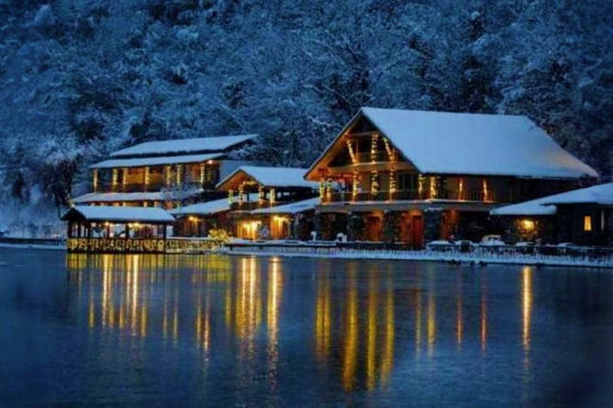 Illuminated log cabin resort at dusk surrounded by snow-covered evergreen forest and frozen lake with golden light reflecting