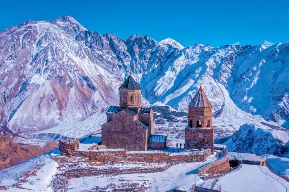 Snow-covered Armenian church with twin bell towers nestled in snow-capped Caucasus mountains under clear blue sky