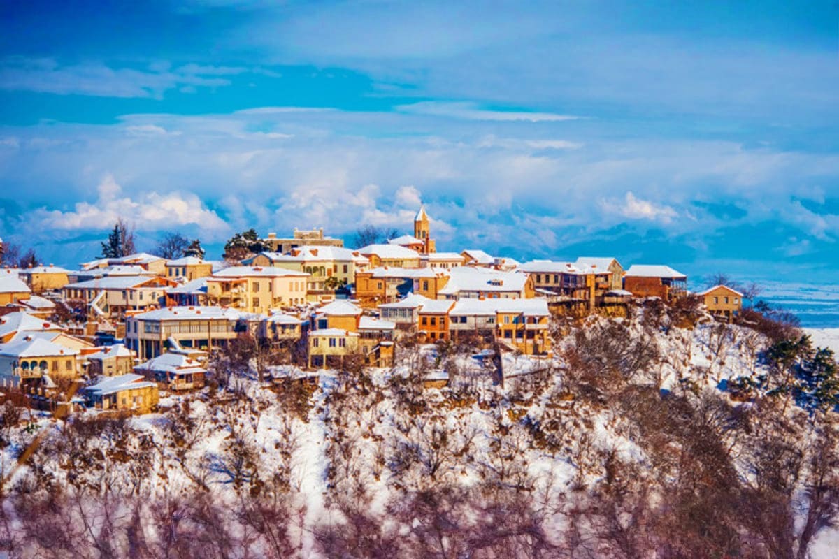 Snow-covered hilltop village with golden-yellow buildings and church spire overlooking winter landscape and mountains in Geor